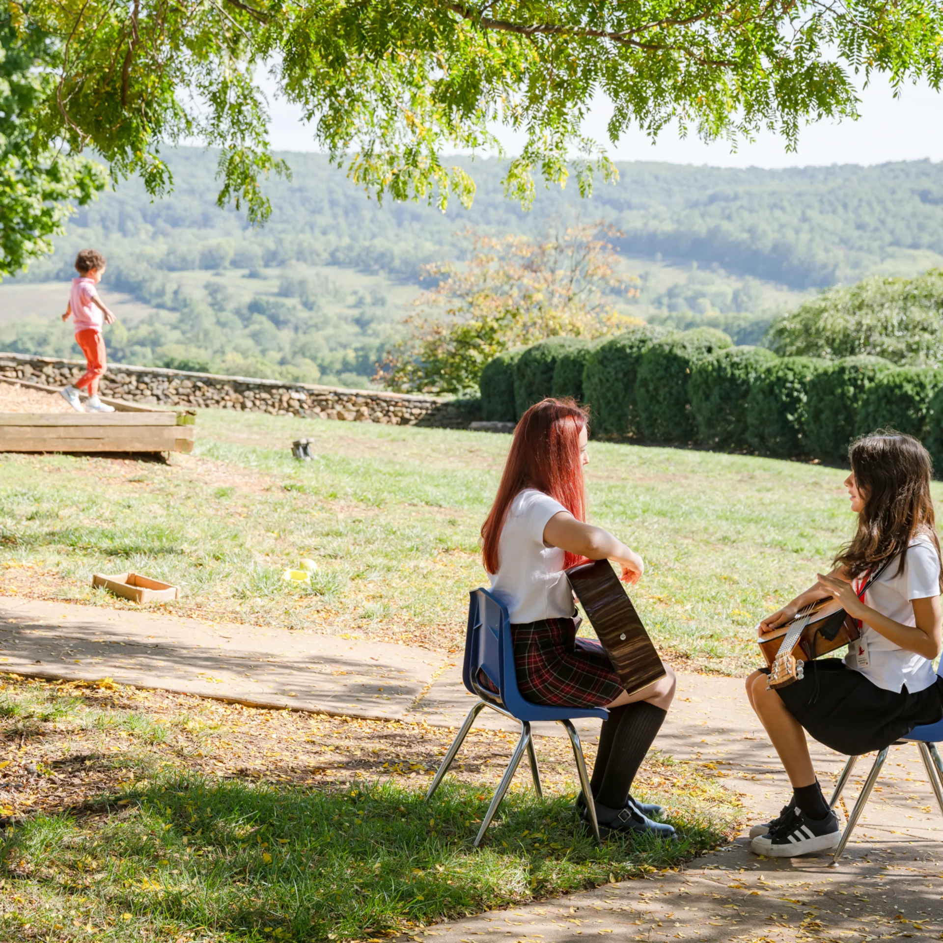students playing guitar outside while younger students play on the playground in the background
